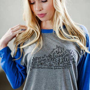 以下が含まれることがあります： グレーとブルーのベースボールTシャツに、黒い文字で「The Bluegrass State」と、ケンタッキー州に関連する単語やフレーズが書かれています。