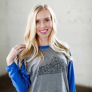 以下が含まれることがあります： グレーとブルーのベースボールTシャツで、黒いグラフィックデザインが施されています。デザインには「Home of the Bluegrass State」という文字と、「VS」、「Hot Brown」、「River」、「Cards」、「Ohio」、「Cats」、「12」、「The Kentucky Derby」、「80」という単語が含まれています。