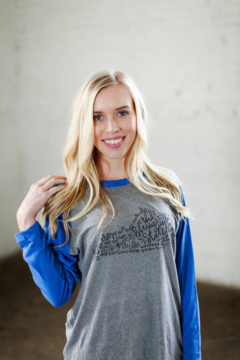 以下が含まれることがあります： グレーとブルーのベースボールTシャツで、黒いグラフィックデザインが施されています。デザインには「Home of the Bluegrass State」という文字と、「VS」、「Hot Brown」、「River」、「Cards」、「Ohio」、「Cats」、「12」、「The Kentucky Derby」、「80」という単語が含まれています。