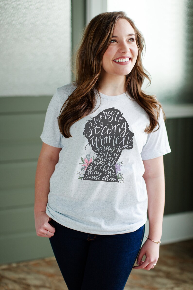 May include: A white t-shirt with a gray silhouette of a woman's head and the text "Here's to strong women. May we know them. May we be them. May we raise them." The silhouette is decorated with pink and purple flowers.