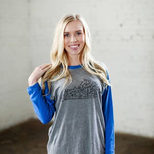 以下が含まれることがあります： グレーとブルーのベースボールTシャツを着た女性。Tシャツには、テネシー州の黒いグラフィックが描かれており、"The Volunteer State"という文字と、テネシー州に関連する他の言葉が書かれています。
