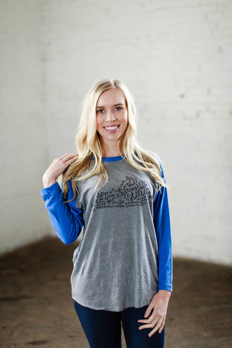 以下が含まれることがあります： グレーとブルーのベースボールTシャツを着た女性。Tシャツには、テネシー州の黒いグラフィックが描かれており、"The Volunteer State"という文字と、テネシー州に関連する他の言葉が書かれています。