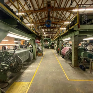 May include: A large industrial space with rows of textile machinery. The machinery is used to weave fabric. The space is well-lit and has a high ceiling.