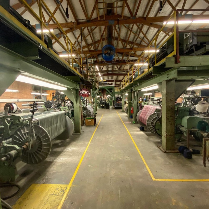 May include: A large industrial space with rows of textile machinery. The machinery is used to weave fabric. The space is well-lit and has a high ceiling.