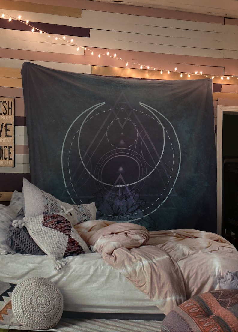 Puede incluir: Un tapiz azul oscuro con un dise&ntilde;o geom&eacute;trico que presenta un c&iacute;rculo, un tri&aacute;ngulo y una flor de loto. El tapiz est&aacute; colgado en una pared con luces de cadena encima. El tapiz est&aacute; en un dormitorio con una cama y almohadas.