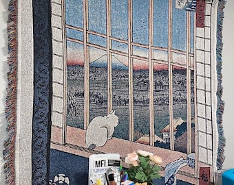 May include: A tapestry wall hanging with a blue and white window scene. The window looks out onto a mountain range and a town. A white cat is sitting on a ledge in the window. The tapestry has a fringe border.