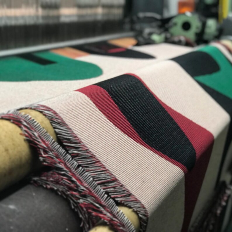 May include: Close-up of a woven fabric with a geometric pattern in black, red, and green. The fabric is being woven on a loom.