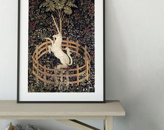 Könnte beinhalten: Gerahmter Wandteppichdruck mit einem weißen Einhorn in einem Holzzaun, umgeben von üppigem Laub und einem Baum. Das Kunstwerk hat einen dunklen Hintergrund mit floralen Details und wird auf einer hellen Holzbank präsentiert.