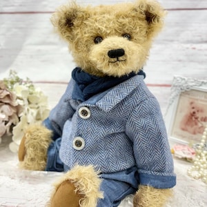 May include: A handcrafted teddy bear with light brown fur, dressed in a blue herringbone jacket and trousers. The bear has a black nose and is seated. The background features a white wooden surface and decorative items.