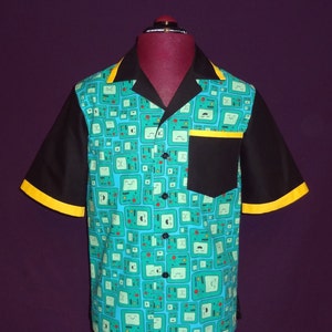 May include: A teal blue short-sleeved button-up shirt with a black collar and black sleeves. The shirt has a black pocket on the left side and a yellow trim on the sleeves. The shirt is covered in a pattern of green and yellow cartoon faces.