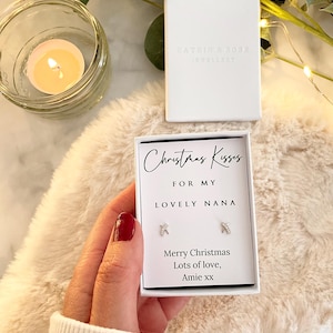 May include: A white gift box with the text "CATKDA ROBR JEWELLERY" and a smaller box with the text "Christmas Kisses FOR MY LOVELY NANA". The smaller box contains a pair of silver earrings. A lit candle is in a glass jar.