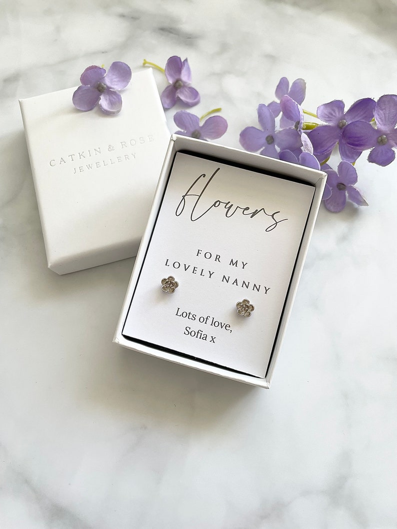 May include: A white gift box with a silver flower earring set inside. The box has a white card with the text "flowers for my lovely nanny lots of love, Sofia x".