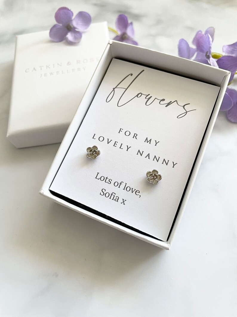 May include: A white gift box with a card that reads "flowers for my lovely nanny" and "lots of love, Sofia x". The box contains a pair of silver flower stud earrings.