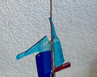 Könnte beinhalten: Ein Mobile aus blauen, roten und weißen Glasstücken, die an einem braunen Draht hängen. Die Glasstücke haben abstrakte Formen und sind in verschiedenen Höhen aufgehängt.