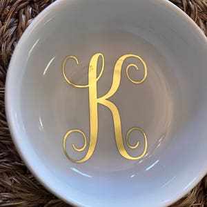 Pode incluir: Um prato de cer&acirc;mica branco com uma letra "K" dourada em it&aacute;lico no centro. O prato tem uma forma curva e est&aacute; sobre uma superf&iacute;cie marrom tecida. A letra dourada &eacute; o ponto focal.