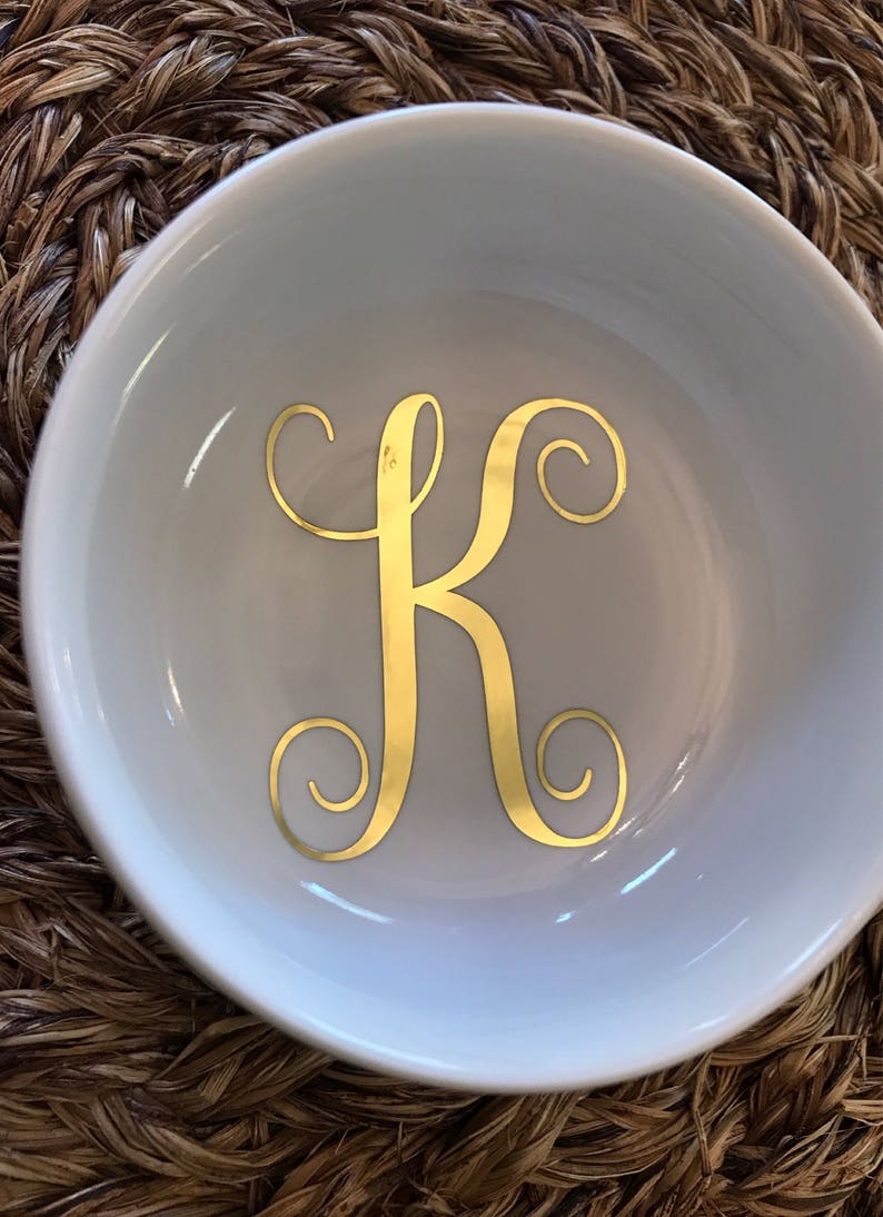 Pode incluir: Um prato de cer&acirc;mica branco com uma letra "K" dourada em it&aacute;lico no centro. O prato tem uma forma curva e est&aacute; sobre uma superf&iacute;cie marrom tecida. A letra dourada &eacute; o ponto focal.