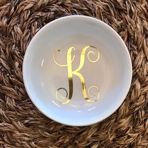 Pode incluir: Uma pequena tigela redonda de cer&acirc;mica branca com uma letra "K" dourada em it&aacute;lico no centro. A tigela est&aacute; sobre uma superf&iacute;cie tecida castanha. A tigela &eacute; provavelmente usada para guardar pequenos objetos, como joias.