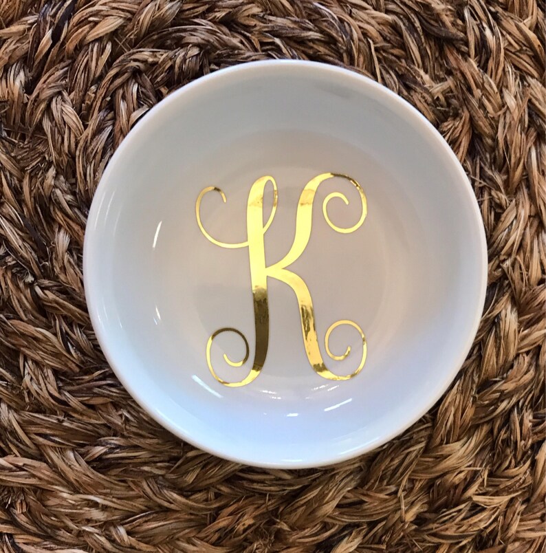 Pode incluir: Uma pequena tigela redonda de cer&acirc;mica branca com uma letra "K" dourada em it&aacute;lico no centro. A tigela est&aacute; sobre uma superf&iacute;cie tecida castanha. A tigela &eacute; provavelmente usada para guardar pequenos objetos, como joias.