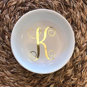 Pode incluir: Uma tigela de cer&acirc;mica branca com a letra "K" dourada em um script decorativo. A tigela est&aacute; centrada em uma superf&iacute;cie tecida marrom. A tigela &eacute; redonda e tem uma borda ligeiramente curvada.