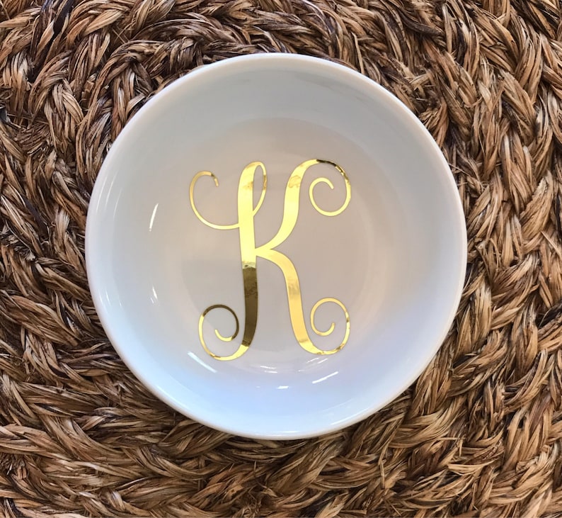 Pode incluir: Uma tigela de cer&acirc;mica branca com a letra "K" dourada em um script decorativo. A tigela est&aacute; centrada em uma superf&iacute;cie tecida marrom. A tigela &eacute; redonda e tem uma borda ligeiramente curvada.