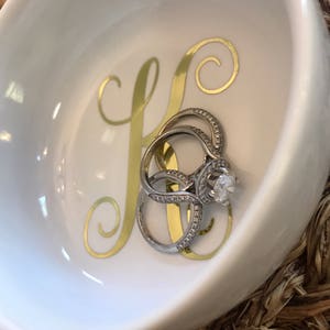 Pode incluir: Uma tigela de cer&acirc;mica branca com uma inicial dourada "K" e um conjunto de an&eacute;is de prata com pedras preciosas transparentes. A tigela tem uma forma curva e est&aacute; sobre uma superf&iacute;cie tecida.