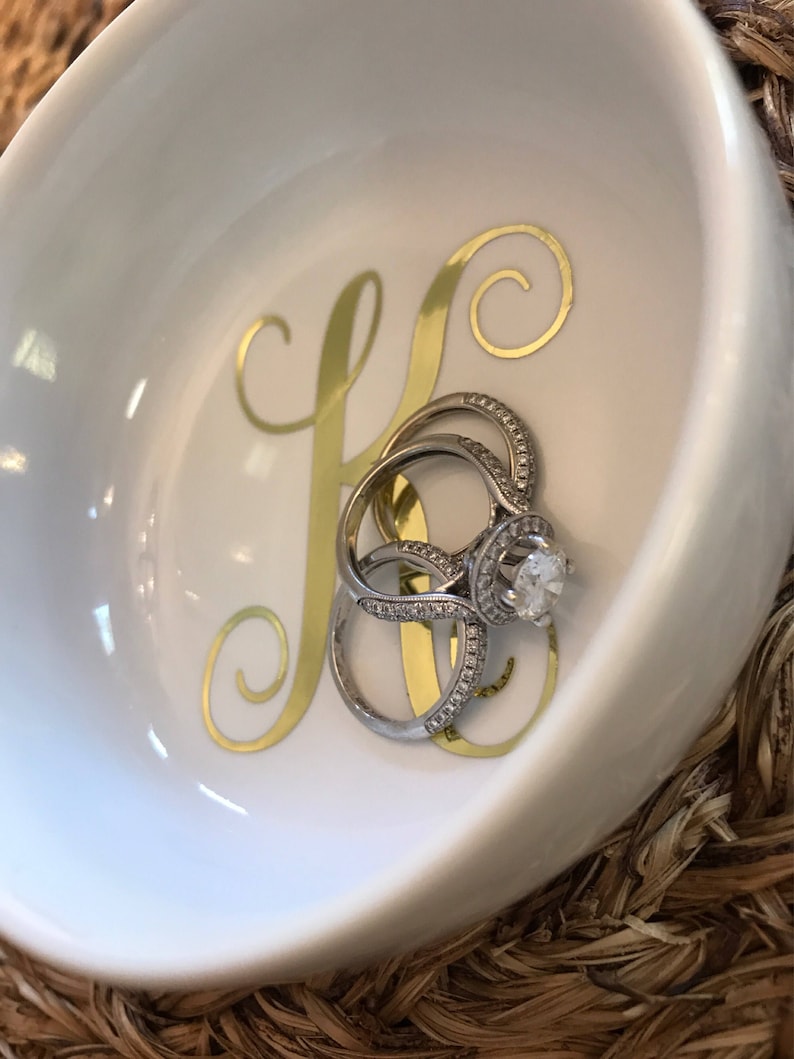 Pode incluir: Uma tigela de cer&acirc;mica branca com uma inicial dourada "K" e um conjunto de an&eacute;is de prata com pedras preciosas transparentes. A tigela tem uma forma curva e est&aacute; sobre uma superf&iacute;cie tecida.