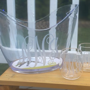 May include: Clear plastic ice bucket and two stemless wine glasses with a monogram design. The bucket has a curved shape with handles. The glasses and bucket are on a wooden surface.