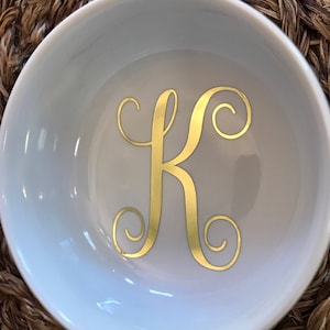 Pode incluir: Uma pequena tigela de cer&acirc;mica branca com a letra "K" dourada em escrita decorativa. A tigela est&aacute; sobre um fundo tecido castanho. O interior da tigela &eacute; liso e refletor.