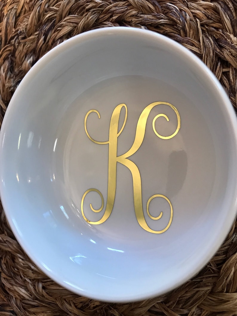 Pode incluir: Uma pequena tigela de cer&acirc;mica branca com a letra "K" dourada em escrita decorativa. A tigela est&aacute; sobre um fundo tecido castanho. O interior da tigela &eacute; liso e refletor.