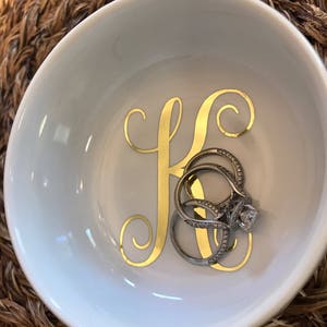 Pode incluir: Uma tigela de cer&acirc;mica branca com um "K" dourado e v&aacute;rios an&eacute;is. Os an&eacute;is s&atilde;o prateados com pedras preciosas transparentes. A tigela &eacute; redonda e est&aacute; sobre uma superf&iacute;cie tecida.