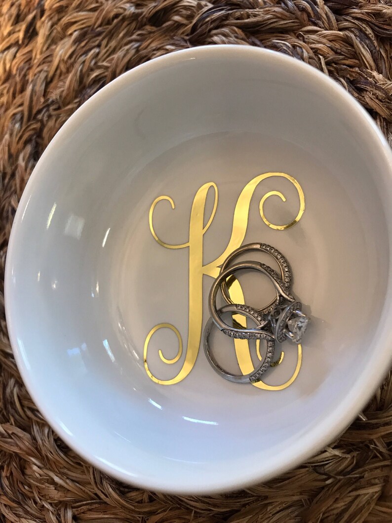 Pode incluir: Uma tigela de cer&acirc;mica branca com um "K" dourado e v&aacute;rios an&eacute;is. Os an&eacute;is s&atilde;o prateados com pedras preciosas transparentes. A tigela &eacute; redonda e est&aacute; sobre uma superf&iacute;cie tecida.
