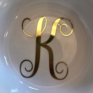 Pode incluir: Tigela de cer&acirc;mica branca com a letra "K" dourada em um script decorativo. A tigela tem um acabamento brilhante e um design simples e elegante. A letra est&aacute; centralizada na tigela.