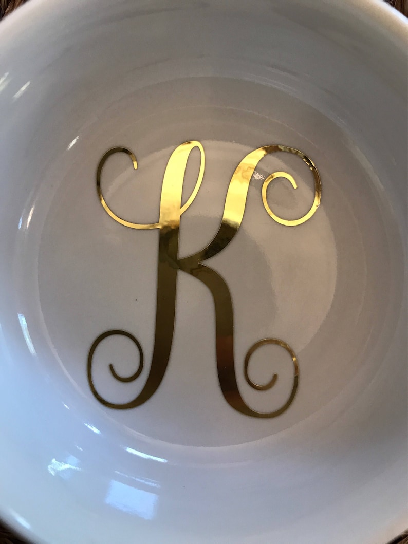 Pode incluir: Tigela de cer&acirc;mica branca com a letra "K" dourada em um script decorativo. A tigela tem um acabamento brilhante e um design simples e elegante. A letra est&aacute; centralizada na tigela.