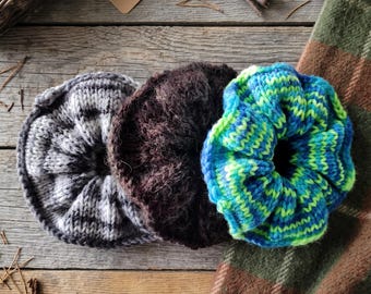 May include: Three circular hair scrunchies made of knitted yarn. One scrunchie is grey and white striped, one is solid dark brown with a fuzzy texture, and one features a vibrant blue and lime green variegated pattern.