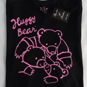 Puede incluir: Camiseta negra con un gráfico rosa de tres osos abrazándose. El texto "Huggy Bear" está impreso sobre los osos. La camiseta tiene una etiqueta que dice "Razens of London".
