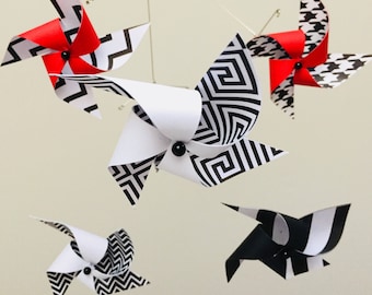 Puede incluir: Un móvil blanco y negro con cinco molinetes de papel. Los molinetes tienen diferentes patrones geométricos, incluyendo zigzags, pata de gallo y un diseño de llave griega. Dos de los molinetes están acentuados con rojo.