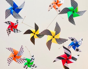 Puede incluir: Un móvil con 10 molinetes coloridos, cada uno con un patrón geométrico diferente. Los molinetes están hechos de papel y tienen detalles en blanco y negro. El móvil está suspendido del techo.