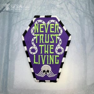 May include: Purple coffin-shaped embroidered patch with the text "NEVER TRUST THE LIVING" in neon green. The patch features a white snake design and a skull with wings. The border is black and white striped. The patch is likely for clothing or accessories.