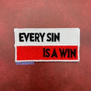 May include: Embroidered patch with the text "EVERY SIN IS A WIN" in black letters. The top half of the patch is white, and the bottom half is red. The patch has a white border.