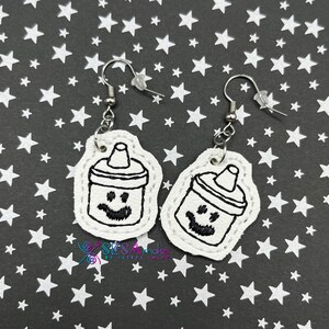 May include: A pair of white leather earrings with a black outline of a baby bottle with a smiling face. The earrings are hanging from silver hooks.