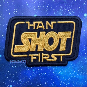 May include: A navy blue embroidered patch with the text "HAN SHOT FIRST" in gold thread. The patch has a rectangular shape with rounded corners and a black border. The text is inside a gold rectangular frame.