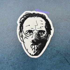 May include: Black and white embroidered patch of a man's face with a mask covering his mouth and nose. The mask has a stitched mouth and a stitched line down the center of the face.