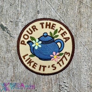 May include: Embroidered patch with a blue teapot design, surrounded by the text "POUR THE TEA LIKE IT'S 1773". The patch features a cream-colored background with a brown border, and floral accents.