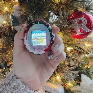 Pode incluir: Um Tamagotchi rosa e branco com uma capa de croch&ecirc; verde e vermelha. A tela mostra uma cena de inverno com tr&ecirc;s personagens. O Tamagotchi est&aacute; sendo segurado por uma m&atilde;o com esmalte de unha colorido.