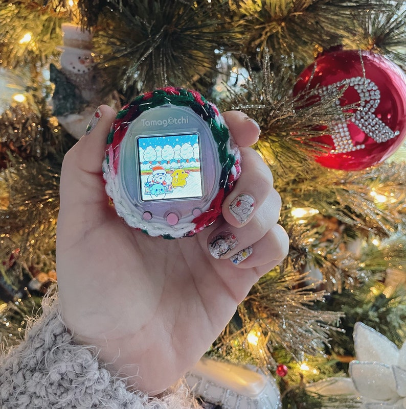 Pode incluir: Um Tamagotchi rosa e branco com uma capa de croch&ecirc; verde e vermelha. A tela mostra uma cena de inverno com tr&ecirc;s personagens. O Tamagotchi est&aacute; sendo segurado por uma m&atilde;o com esmalte de unha colorido.