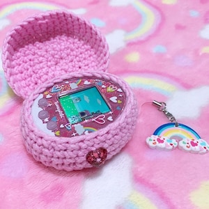 Pode incluir: Uma capa de croch&ecirc; rosa com um Tamagotchi rosa e branco dentro. O Tamagotchi tem um design de arco-&iacute;ris e cora&ccedil;&otilde;es. Um amuleto de arco-&iacute;ris rosa e branco est&aacute; preso &agrave; capa.