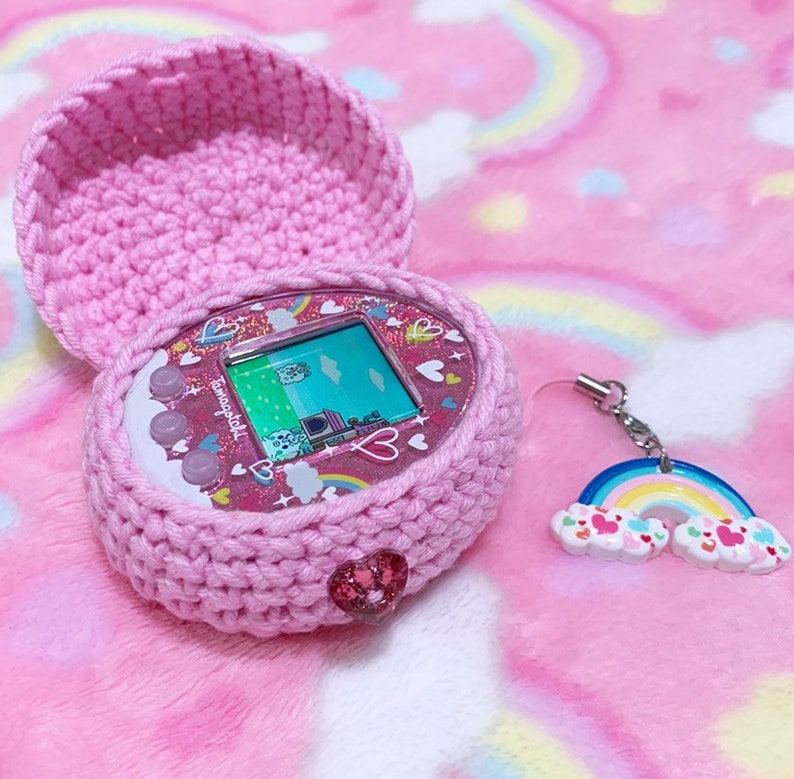 Pode incluir: Uma capa de croch&ecirc; rosa com um Tamagotchi rosa e branco dentro. O Tamagotchi tem um design de arco-&iacute;ris e cora&ccedil;&otilde;es. Um amuleto de arco-&iacute;ris rosa e branco est&aacute; preso &agrave; capa.