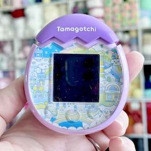 Peut inclure: Jouet Tamagotchi violet avec un motif arc-en-ciel et nuage. L'écran est noir et le jouet porte l'inscription "Tamagotchi" en haut.