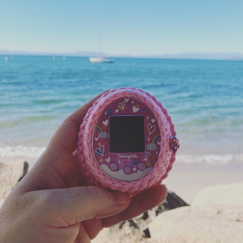 Pode incluir: Brinquedo Tamagotchi rosa com um design de arco-&iacute;ris e cora&ccedil;&atilde;o, em uma capa de croch&ecirc; rosa. O brinquedo est&aacute; sendo segurado em uma m&atilde;o contra um fundo de praia e oceano.