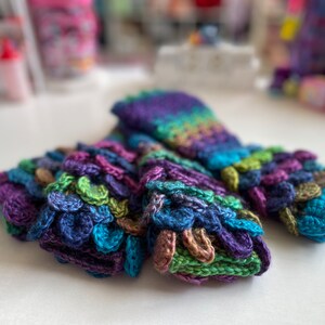 May include: Three pairs of colorful crocheted fingerless gloves with a textured, scaly pattern. The gloves are made in shades of purple, blue, green, and brown.